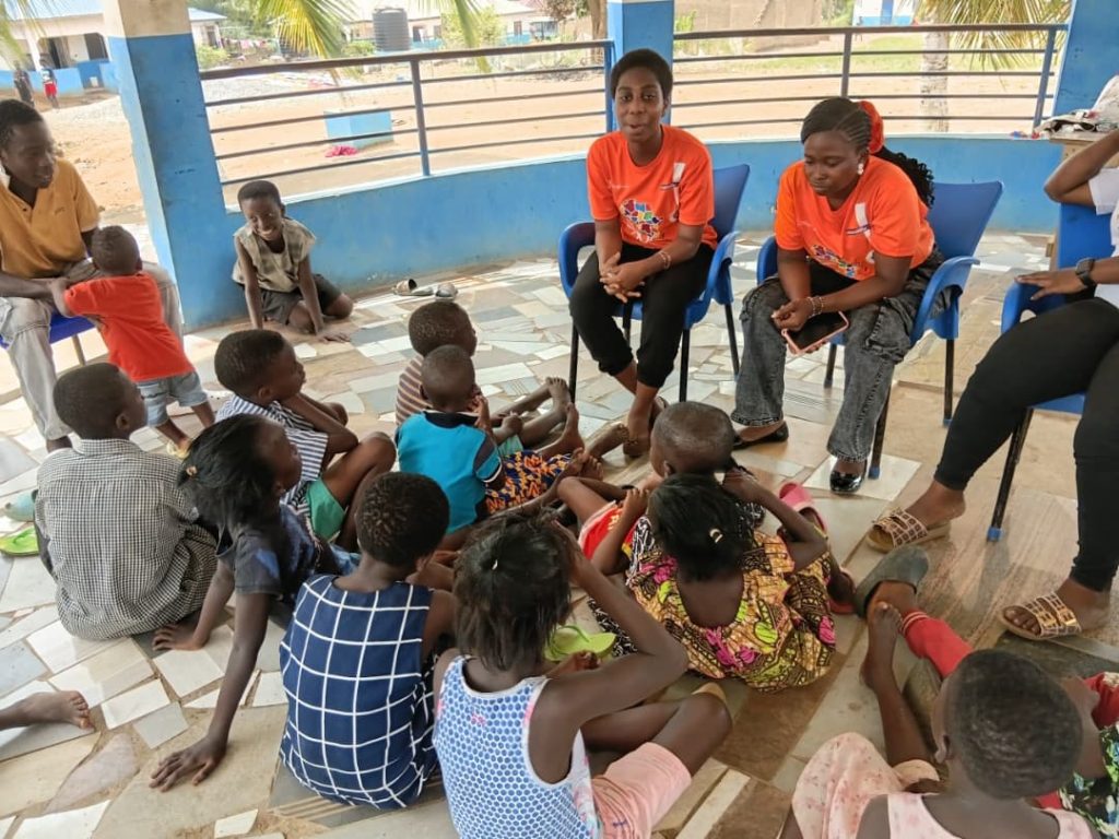 Volunteers in orange shirts sit with children during a community visit.